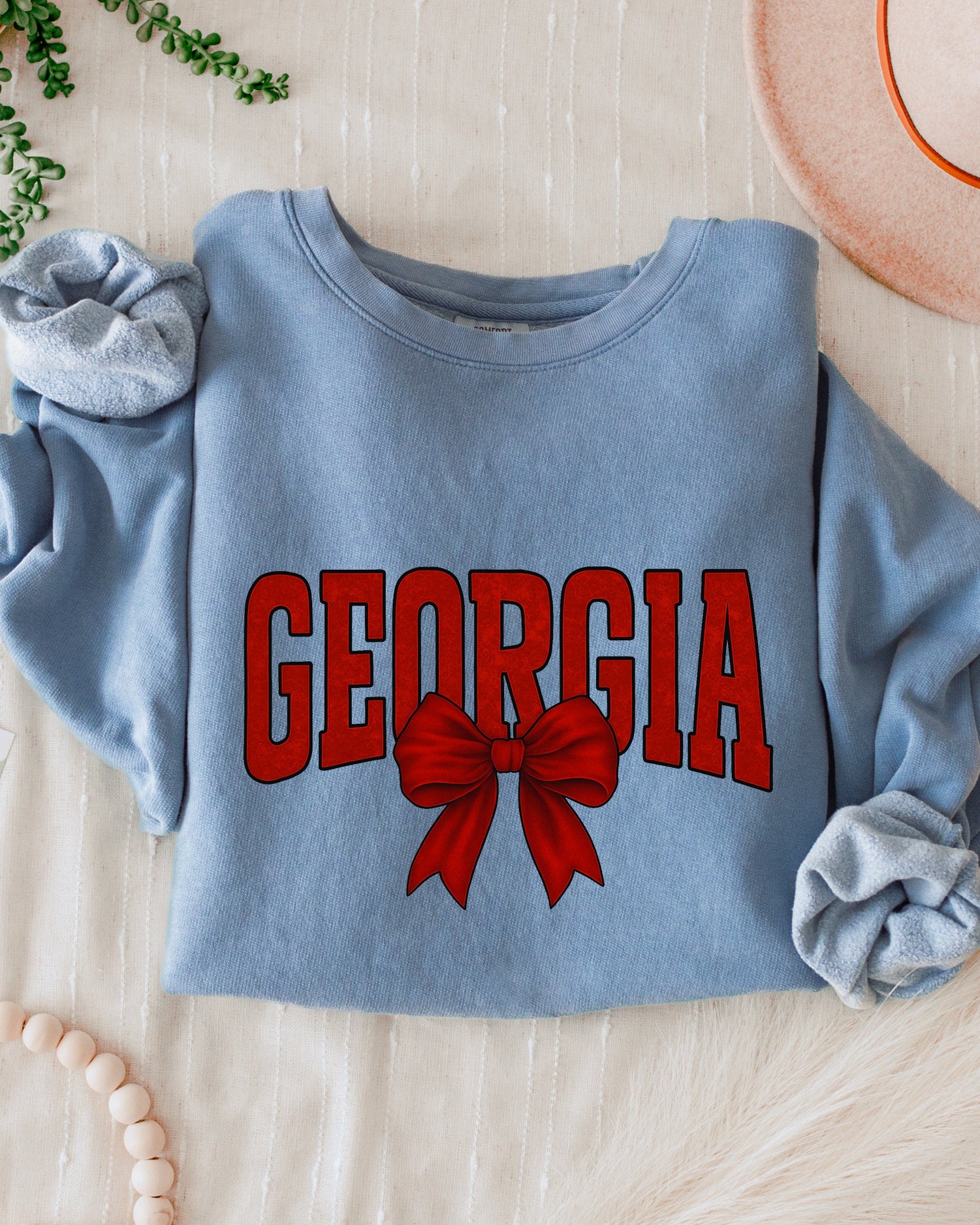 Blue sweatshirt with 'Georgia' and a bow design on a wooden surface with accessories.