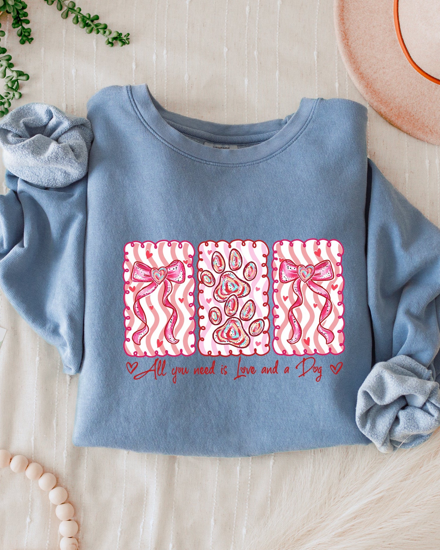 Blue sweatshirt with pink designs and text, accompanied by scrunchies and a hat on a light wooden surface.