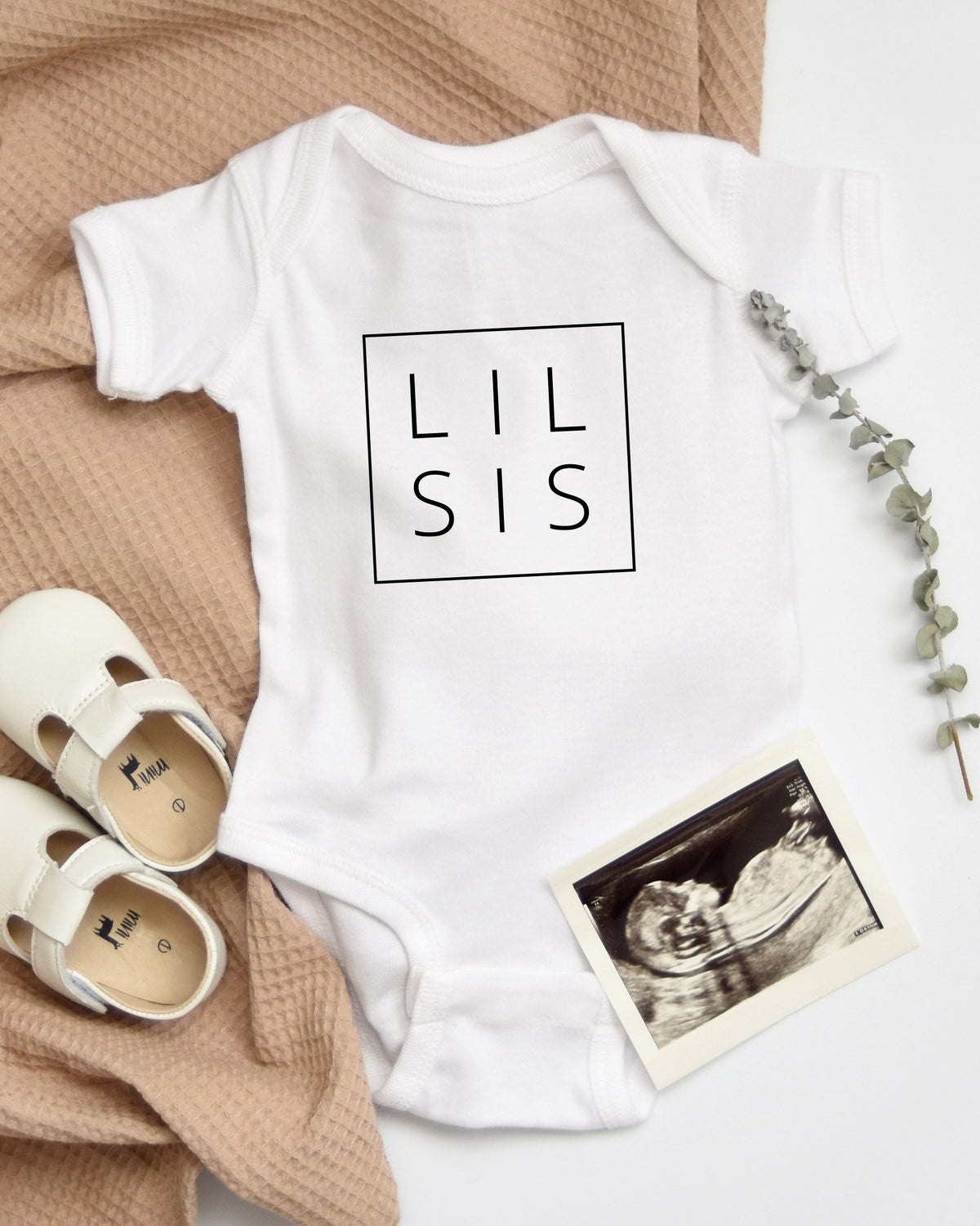 White cotton "LIL SIS" baby onesie with envelope neck, snap leg closure, white baby shoes, and sonogram.