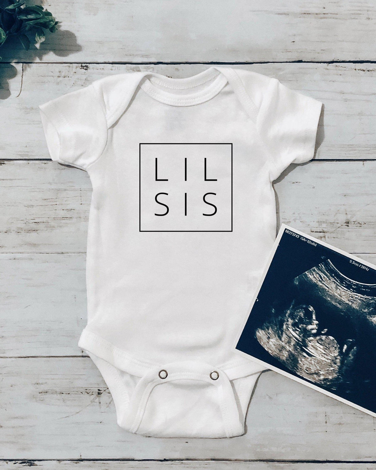 White cotton baby onesie with "LIL SIS" in a black box, next to an ultrasound image.