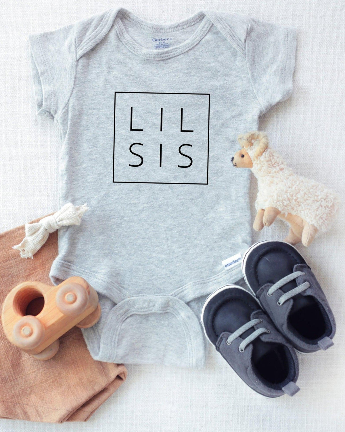 Light gray cotton baby onesie with "LIL SIS" in a black box, displayed with baby shoes and toys.