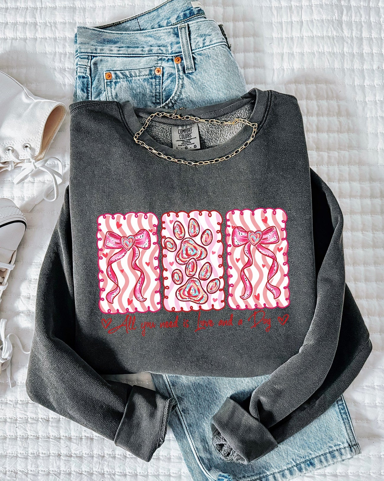Gray sweatshirt with pink designs and text, white sneakers, and blue jeans on a textured surface